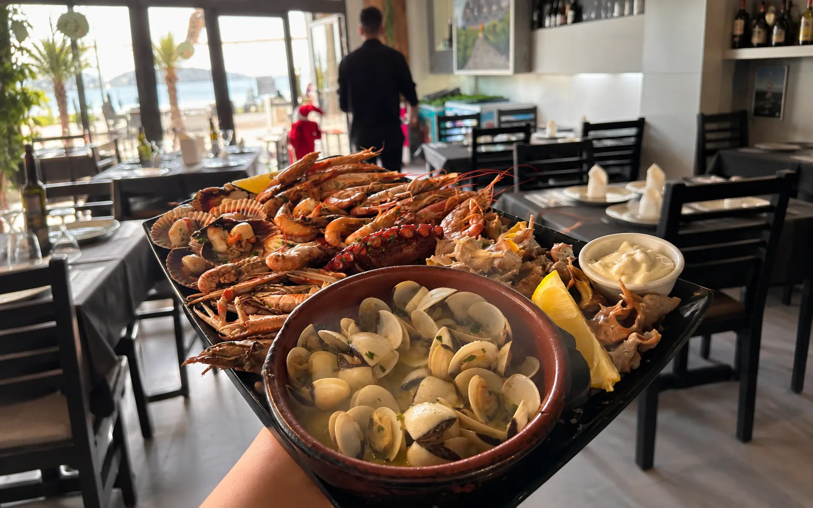 Bandeja de marisco a la brasa con pulpo, langostinos y cigalas, y una cazuela de almejas en primer plano, en la sala de El Poal